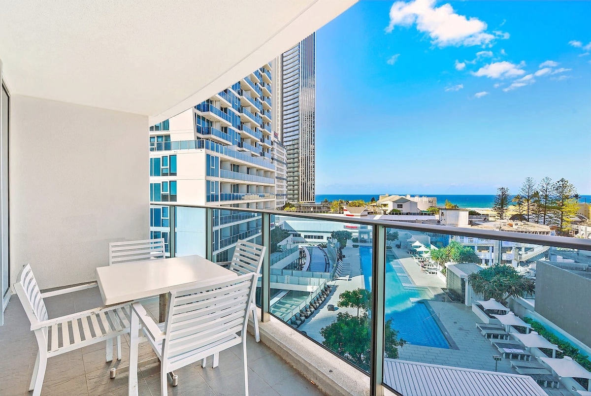 A spacious balcony is equipped with a white outdoor dining set featuring a rectangular table and six chairs. Expansive views of the ocean and surrounding buildings are visible through the glass railing, complemented by blue skies and wispy clouds.