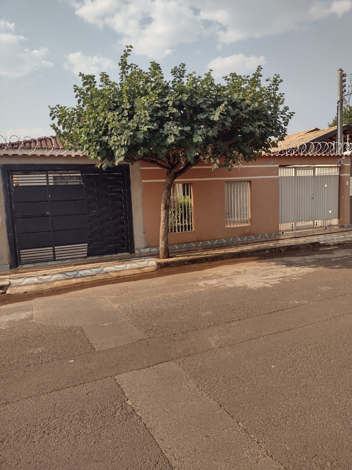 A street view of the property is captured, showcasing a sturdy gated entrance framed by a leafy tree. The building's exterior features a neutral colour with vertical bars on the windows, providing a sense of privacy. The well-maintained sidewalk leads towards the entrance.