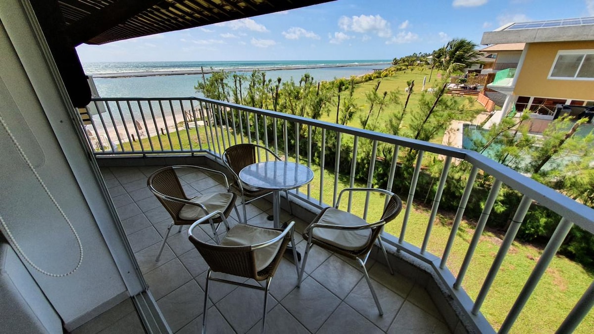 A balcony area features a round table surrounded by four chairs, offering a view of the expansive ocean and coastline. The scene is complemented by lush green grass and palm trees in the foreground, with a clear sky and sunlight enhancing the atmosphere.