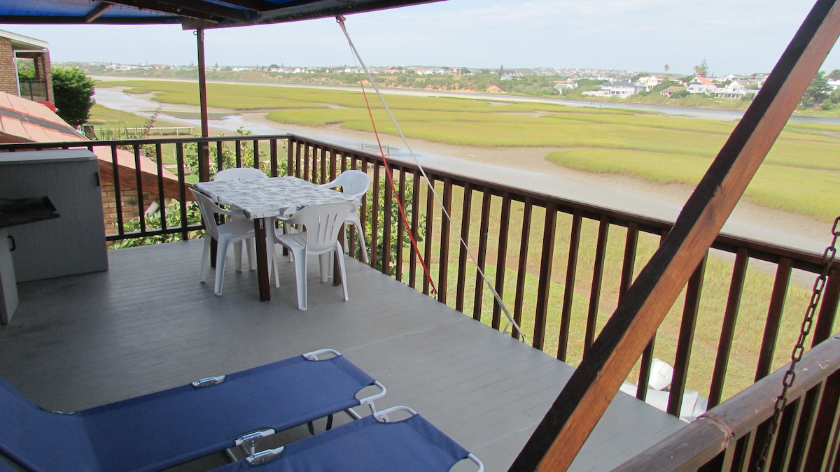 A deck area features a dining table surrounded by white chairs, offering a scenic view of the tidal river and surrounding landscape. Two lounge chairs are positioned for relaxation. The open space is shaded by a blue canopy, enhancing comfort while enjoying the outdoors.