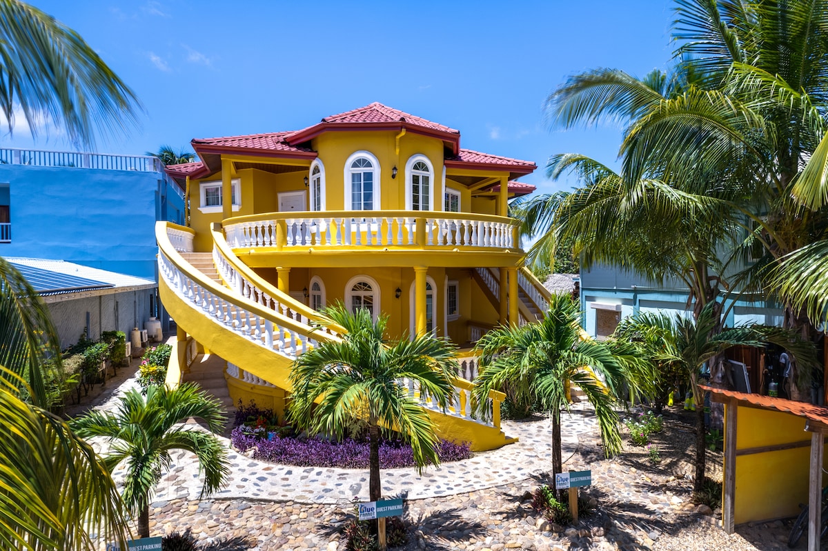 A vibrant yellow beachfront home is surrounded by tropical palm trees. The two-story structure features a distinctive spiral staircase leading to a spacious balcony. Large arched windows allow natural light to fill the interior. The pathway is lined with decorative stones and lush, flowering plants.