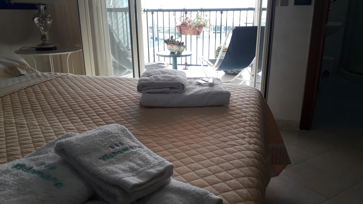 A neatly made spacious bed is adorned with a quilted coverlet. Fresh towels are arranged on the bed. Behind, a balcony is visible with potted plants and a small table set for two, offering a glimpse of the view outside.