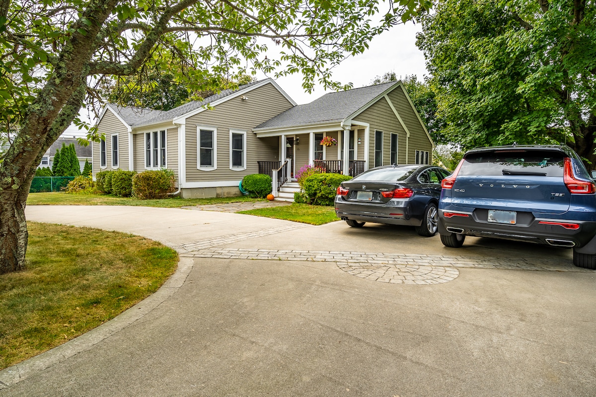 A single-story beach house is set in a residential area, featuring a circular driveway that accommodates several vehicles. Lush greenery and bushes surround the home, while a large tree provides shade near the front entrance.