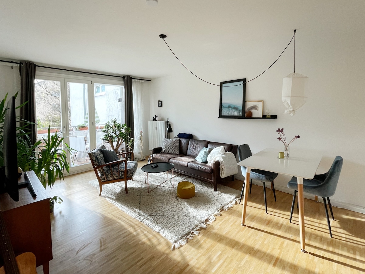 A bright living room features large glass doors leading to a balcony, allowing natural light to flood the space. A cozy seating area includes a leather sofa and a pair of armchairs, while a dining table with chairs invites meals together. A soft area rug adds warmth.