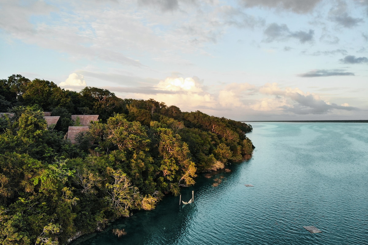 A tranquil view of the azure lagoon is framed by lush greenery and trees. Several rustic cabins can be seen nestled among the foliage, their roofs blending harmoniously with the natural surroundings. The calm water reflects the soft colors of the sky during sunset.
