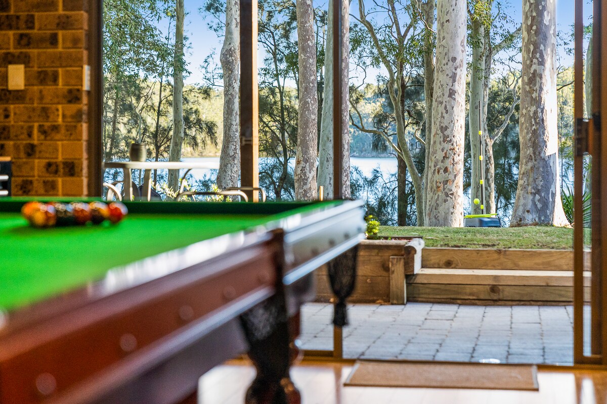 A well-lit games room features a green felt pool table in the foreground, with large glass doors opening to a scenic lakeside view. Tall trees frame the outdoor area, creating a natural and inviting atmosphere.