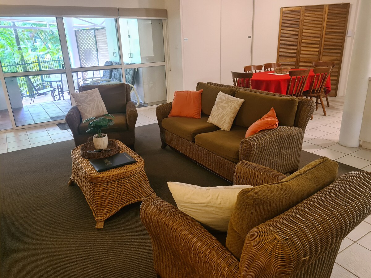 A comfortable living area is presented with seating on two wicker sofas, complemented by cushions in various shades. A coffee table is centered among the seating, while a dining table is visible in the background, accommodating multiple chairs. Natural light enters through large sliding glass doors.