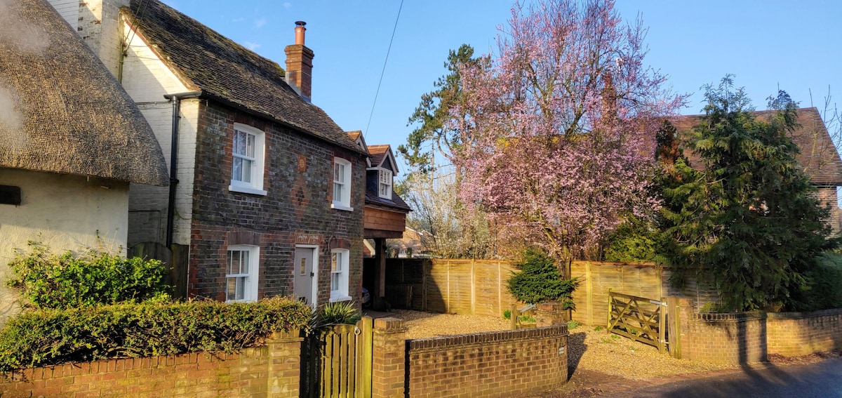 A charming cottage with a thatched roof is set against a clear blue sky. Surrounding greenery includes well-maintained hedges and blooming trees in shades of pink. The property features white windows and a gravel pathway leading to the entrance, blending harmoniously into the tranquil environment.