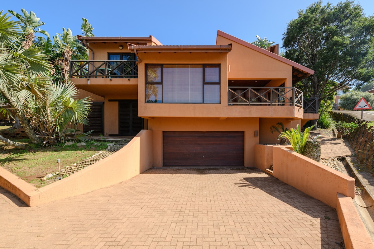 A two-story orange-colored house features a spacious front driveway and a large garage door. A wrap-around balcony is visible on the upper level, surrounded by greenery and distant trees. Sunlight brightens the exterior against a clear blue sky.