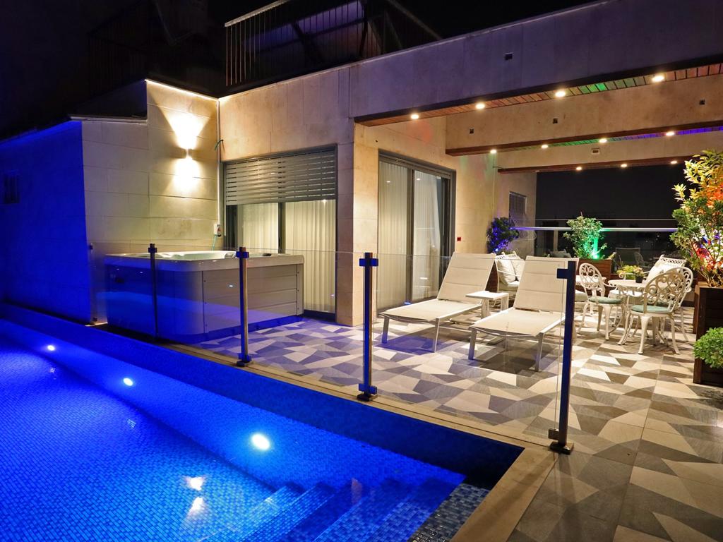 The image captures a beautifully designed terrace featuring a private swimming pool illuminated in blue lighting. A jacuzzi is positioned nearby, accompanied by loungers and a dining area adorned with greenery. Large sliding doors offer access to the indoor space, enhancing the inviting atmosphere.