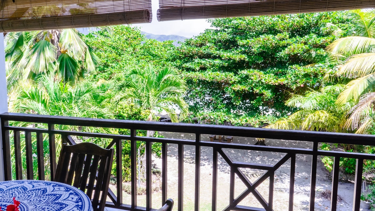 A wooden balcony with decorative railings offers a view of lush greenery and palm trees. A circular blue-patterned table is positioned against the railing, complementing the natural landscape visible beyond.