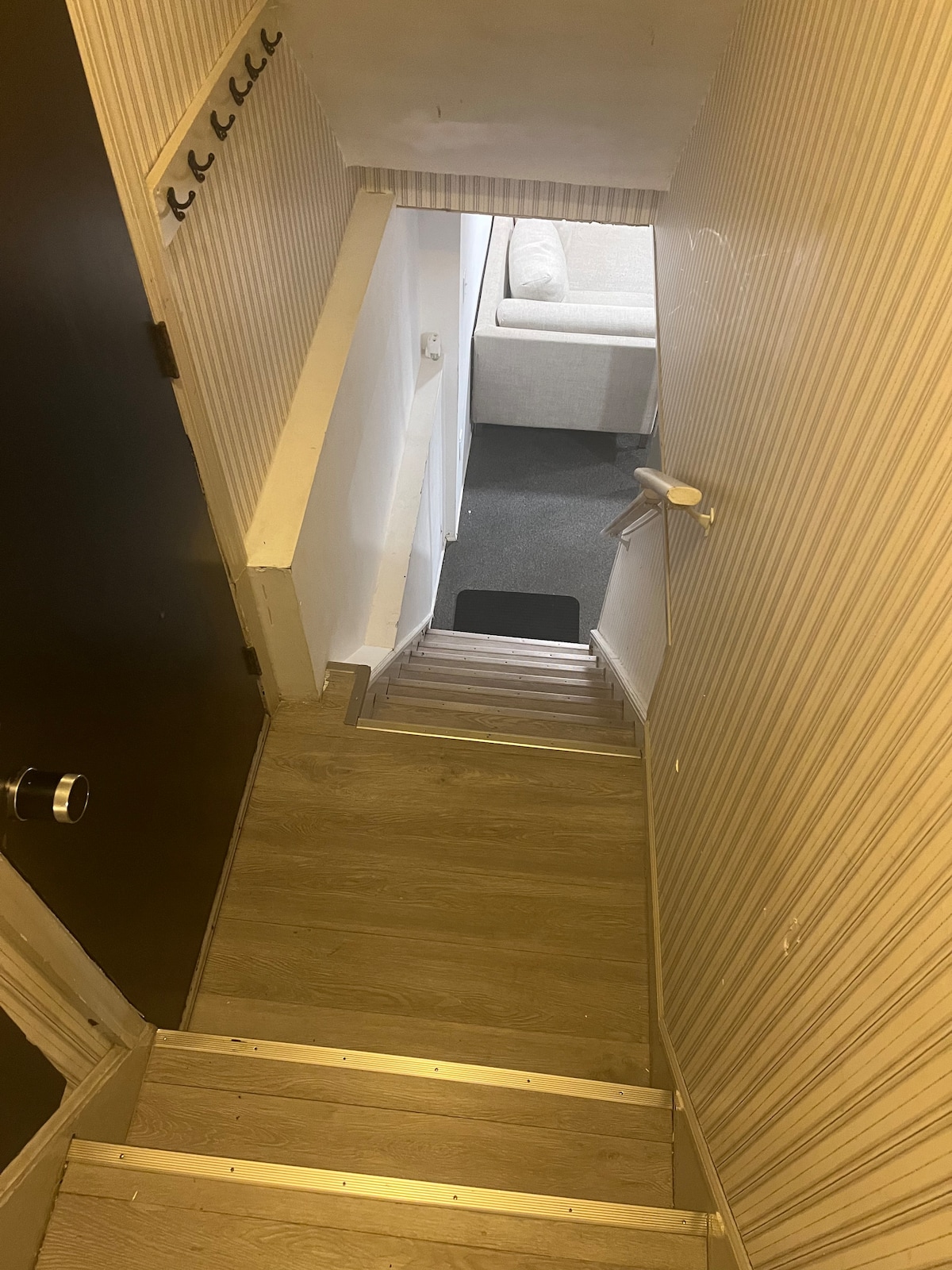 A staircase leads downwards, featuring wooden steps and a railing. The walls are covered in subtle striped wallpaper. At the bottom, a glimpse of the living area is visible, showcasing a light-colored sectional sofa.