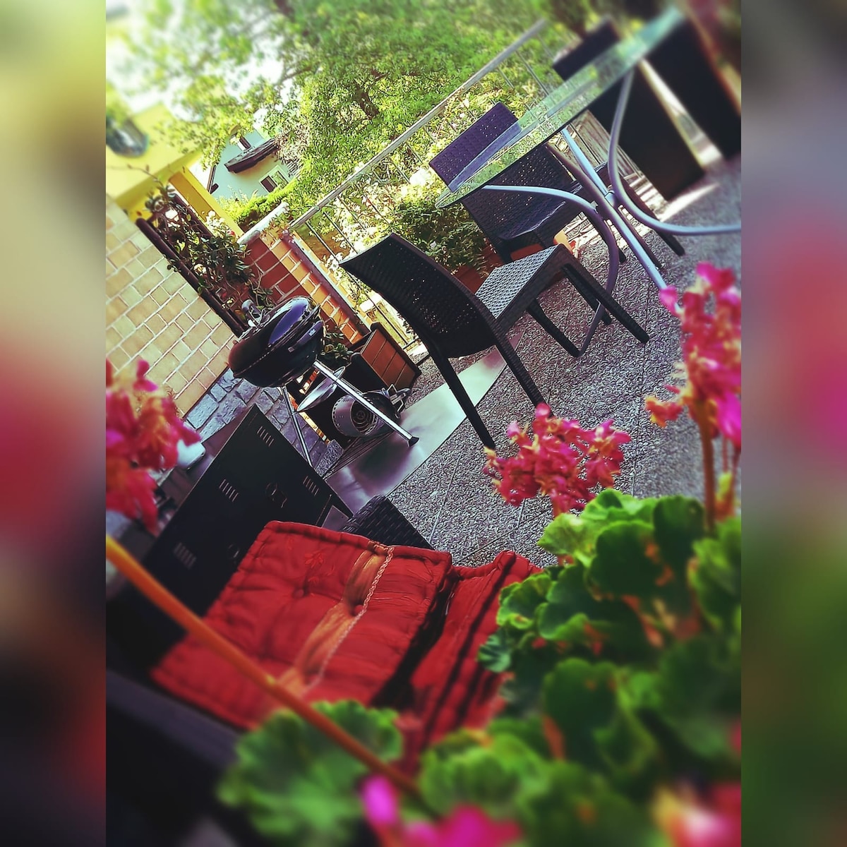 A terrace area is furnished with a dining table and chairs, accompanied by a barbecue grill. Colorful flowers in the foreground add vibrancy to the space, creating a pleasant outdoor setting surrounded by greenery.