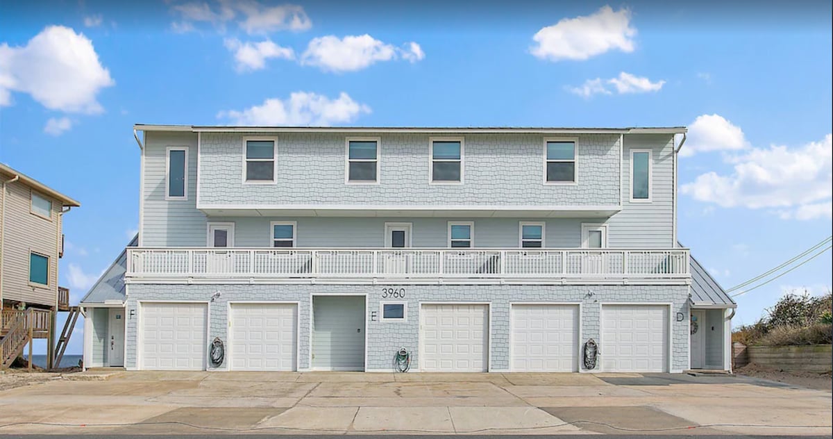 A three-story beachfront townhome is presented with a light blue exterior. Balconies extend across the front, and multiple garage doors are visible at ground level. The sky is bright with scattered clouds, enhancing the home's inviting seaside appeal.