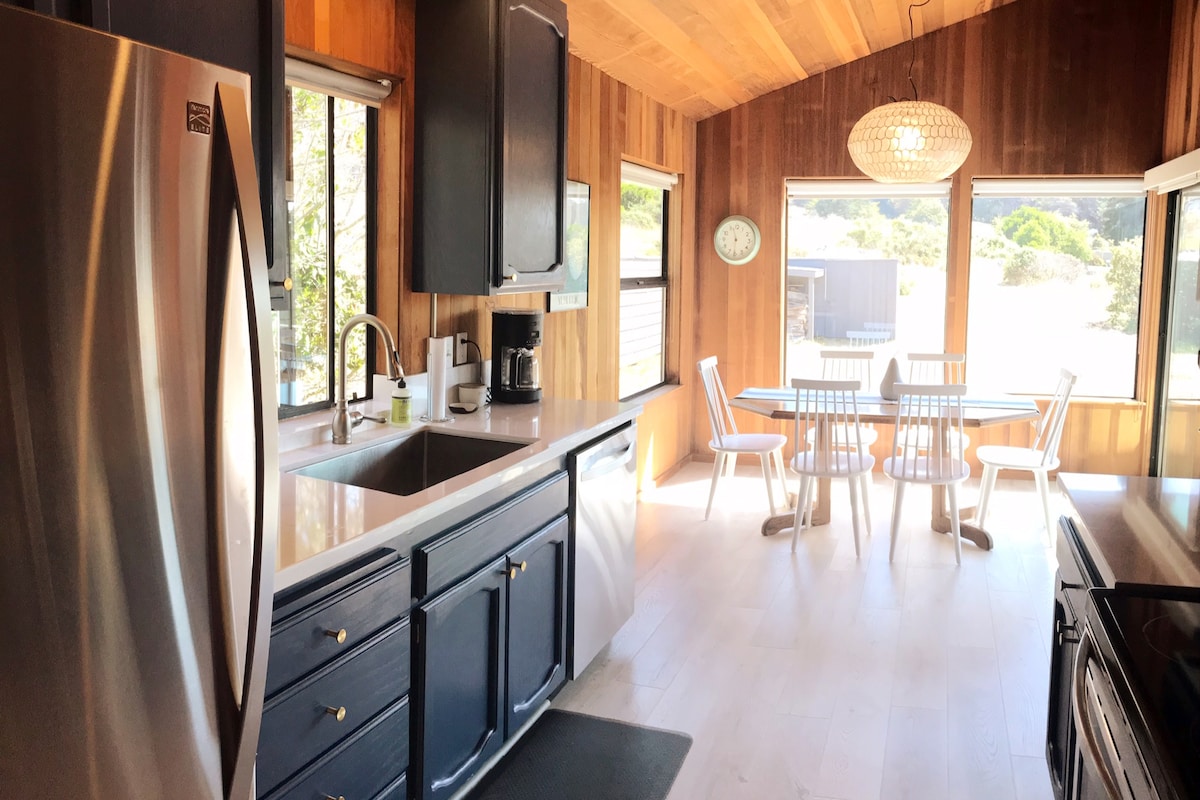 An updated kitchen area features dark cabinetry and stainless steel appliances, with ample counter space around a sink. Natural light enters through large windows, illuminating a dining table with white chairs. Wood paneling adds warmth to the space, creating a welcoming atmosphere.