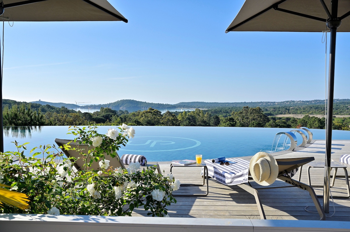An infinity pool stretches towards a scenic landscape, framed by blooming white flowers. Loungers are positioned on the wooden deck, accompanied by a small table adorned with refreshments. Sun umbrellas provide shade, enhancing the tranquil setting with views of rolling hills and a distant body of water.