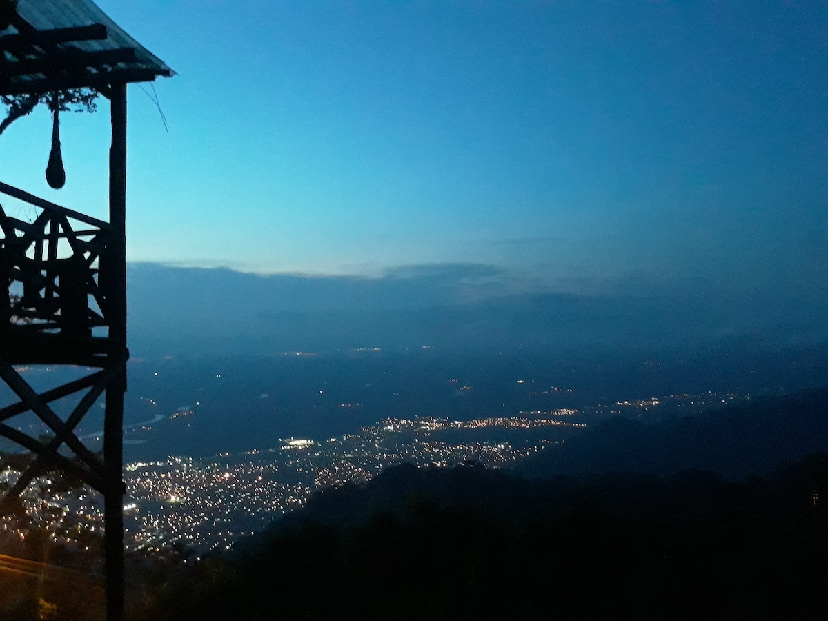 A panoramic view showcases a valley illuminated by city lights under a twilight sky. The silhouette of a wooden structure is visible in the foreground, adding depth to the sweeping landscape. The colors transition from deep blue to soft, muted hues as daylight fades.