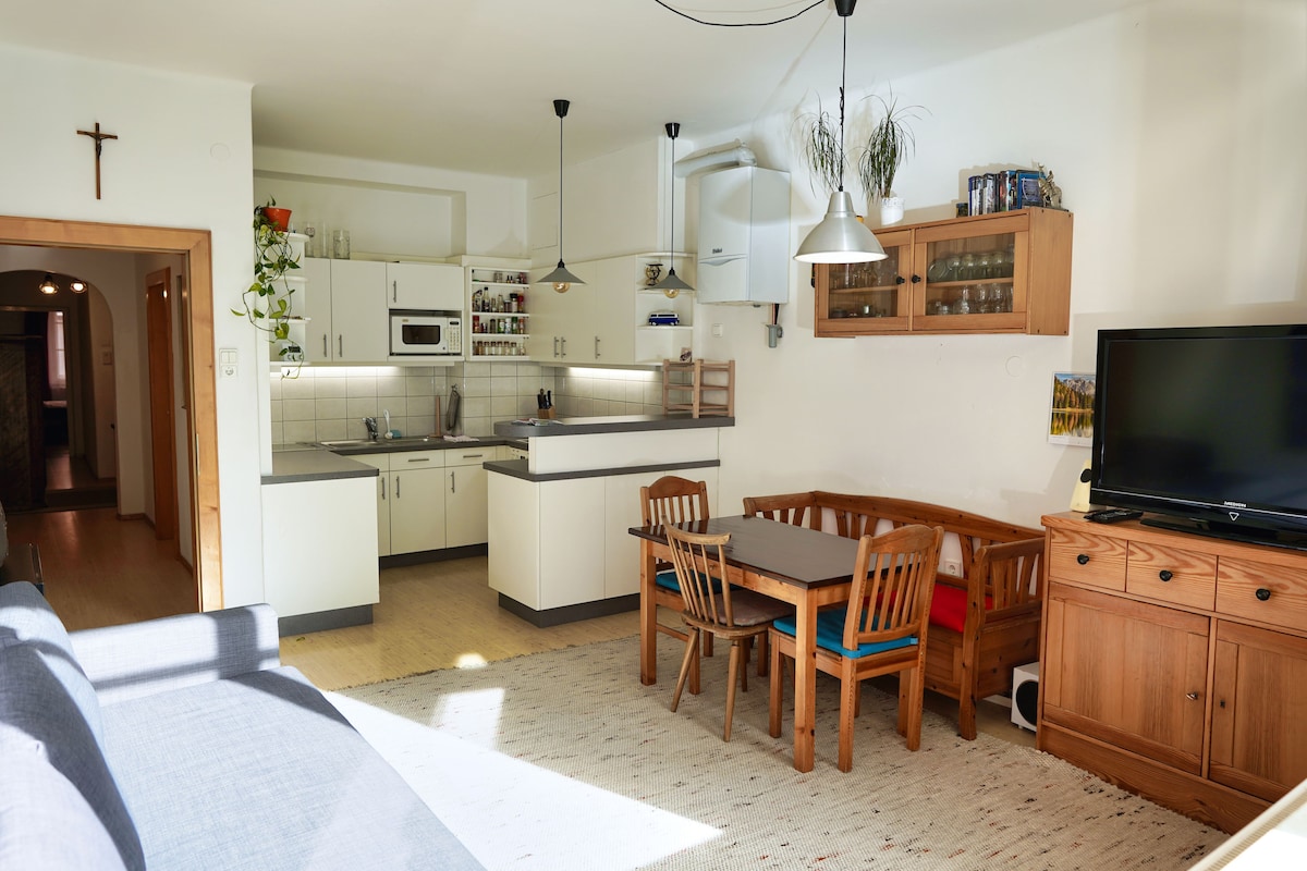 An open kitchen and living area are presented, featuring white cabinetry and various appliances, including a microwave and stove. A dining table with four wooden chairs sits adjacent to a light brown sideboard. Natural light illuminates the space, complementing the neutral-toned floor and decor.