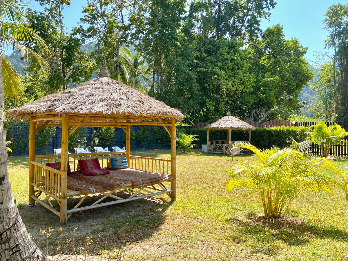 A spacious outdoor area is showcased with two bamboo salas surrounded by lush greenery. Comfortable seating is provided on the larger sala, adorned with colorful cushions. The scene reflects a tranquil environment with sunlight filtering through the surrounding trees, inviting relaxation.