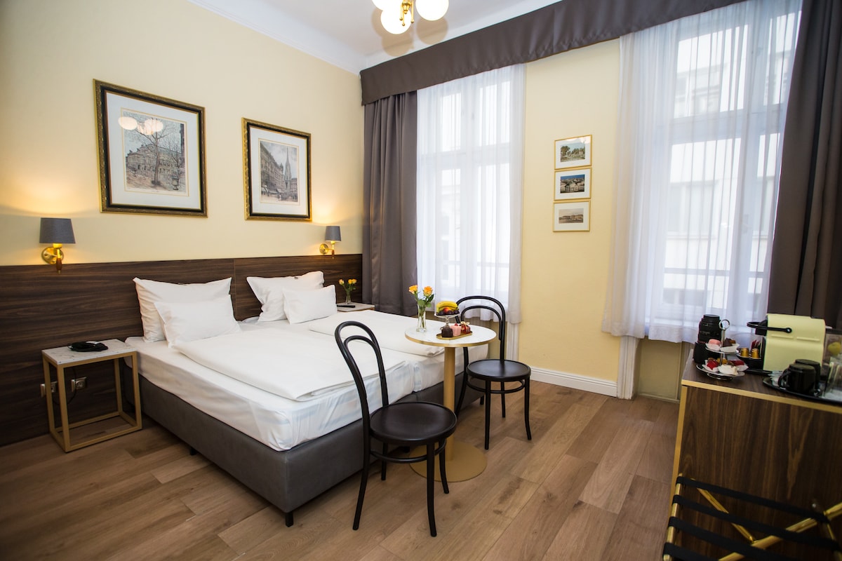 A guest room features a spacious bed with white linens, framed artwork on the walls, and large windows allowing natural light. A small round table with two chairs is positioned beside the bed, showcasing a simple decorative arrangement. A desk in the corner adds functionality.