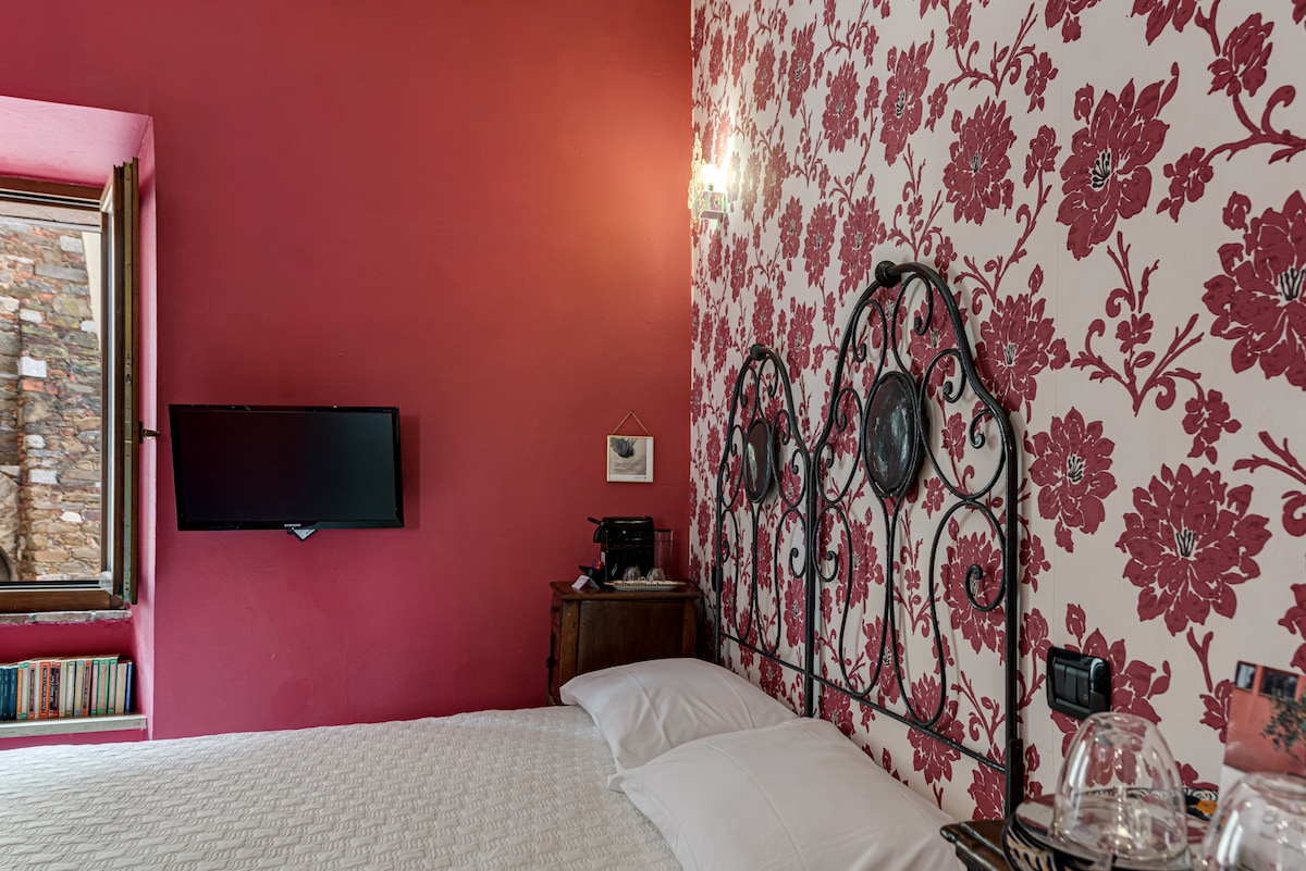 A charming bedroom features a decorative floral wallpaper in deep red tones. A wrought-iron bed rests against the wall, and a flat-screen TV is mounted nearby. Natural light enters through a window, illuminating the space, while a vintage nightstand adds character.