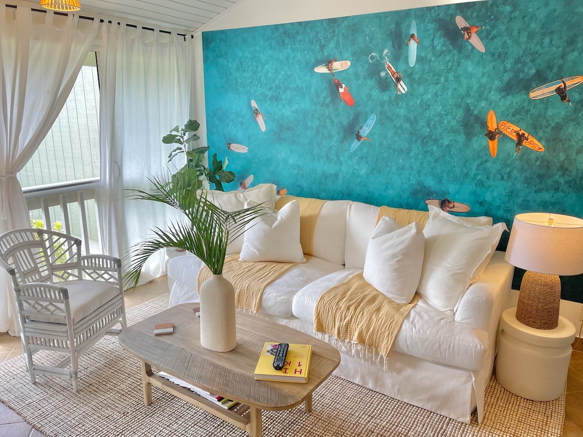 A bright living area is accented by a large wall mural depicting surfboards on turquoise waters. A comfortable white sofa is adorned with plush pillows and a soft throw. A natural fiber rug covers the floor, complemented by a round lamp and a stylish coffee table.