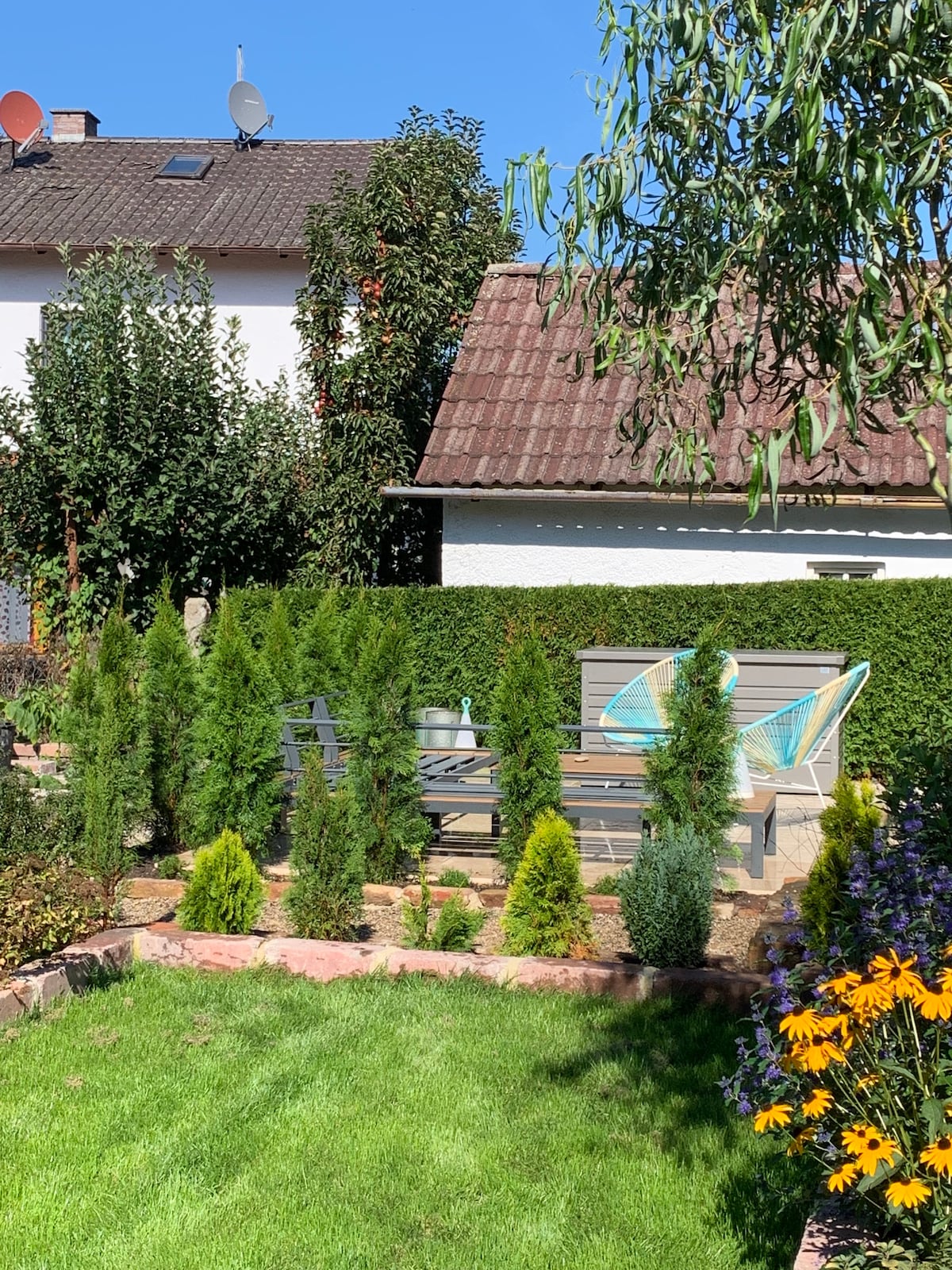 A serene garden area is highlighted, featuring neatly trimmed hedges and a lush green lawn. Two comfortable chairs are positioned nearby, accompanied by a small table. Seasonal flowers add color, with a backdrop of a rustic house and clear blue skies above.