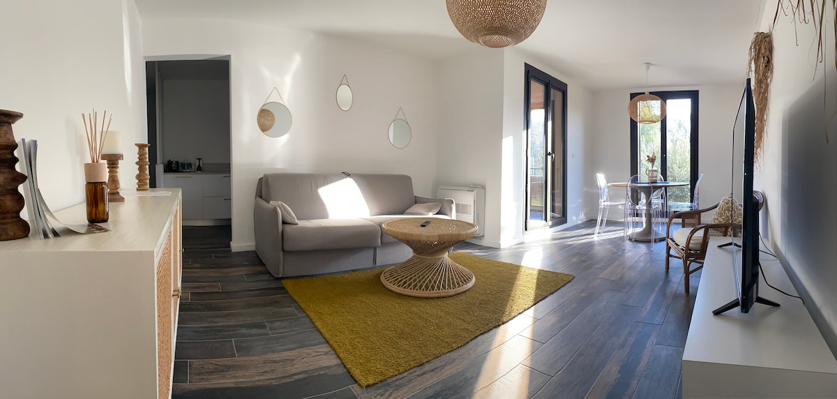 A bright living area features a spacious sofa against a light wall, complemented by a round coffee table on an area rug. Large glass doors provide access to the outdoor space, while woven decorative elements add texture. Natural light enhances the refreshing ambiance.