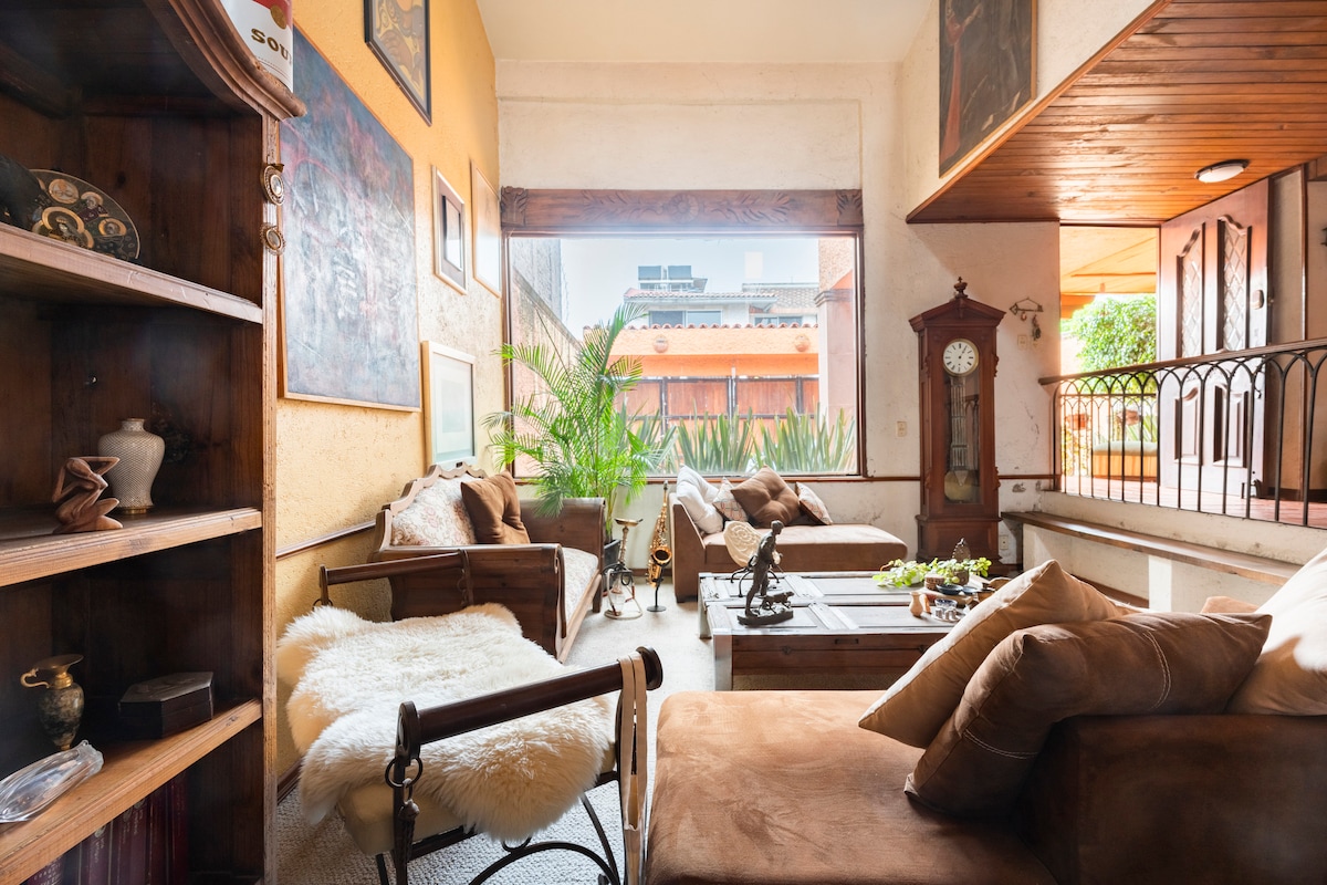 A spacious living area features comfortable seating arranged around a wooden coffee table. Large windows provide natural light and views of the greenery outside. Decorative art pieces adorn the walls, adding character to the cozy ambiance.