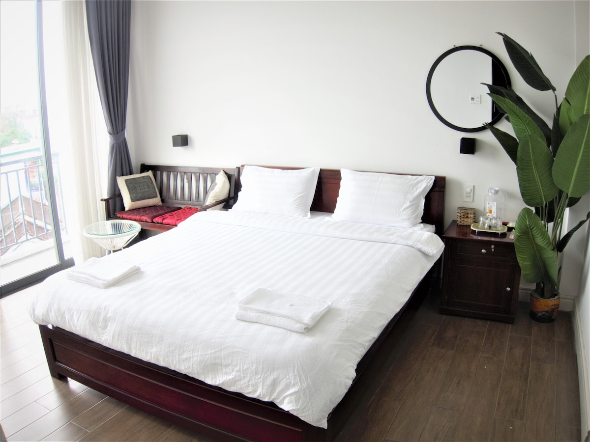 A well-lit bedroom features a spacious bed with crisp white linens. A comfortable seating area can be seen beside the bed, along with a round mirror mounted on the wall. A large plant adds a touch of greenery, and the balcony door opens to a view.