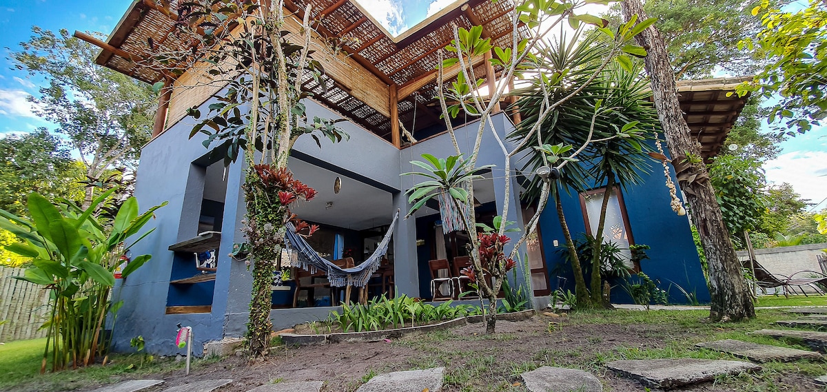 The image showcases a spacious house surrounded by a lush garden. The exterior features a mix of wooden and blue walls, with a hammock set among tropical plants. Seating areas and open spaces invite relaxation under natural light, creating a welcoming atmosphere.
