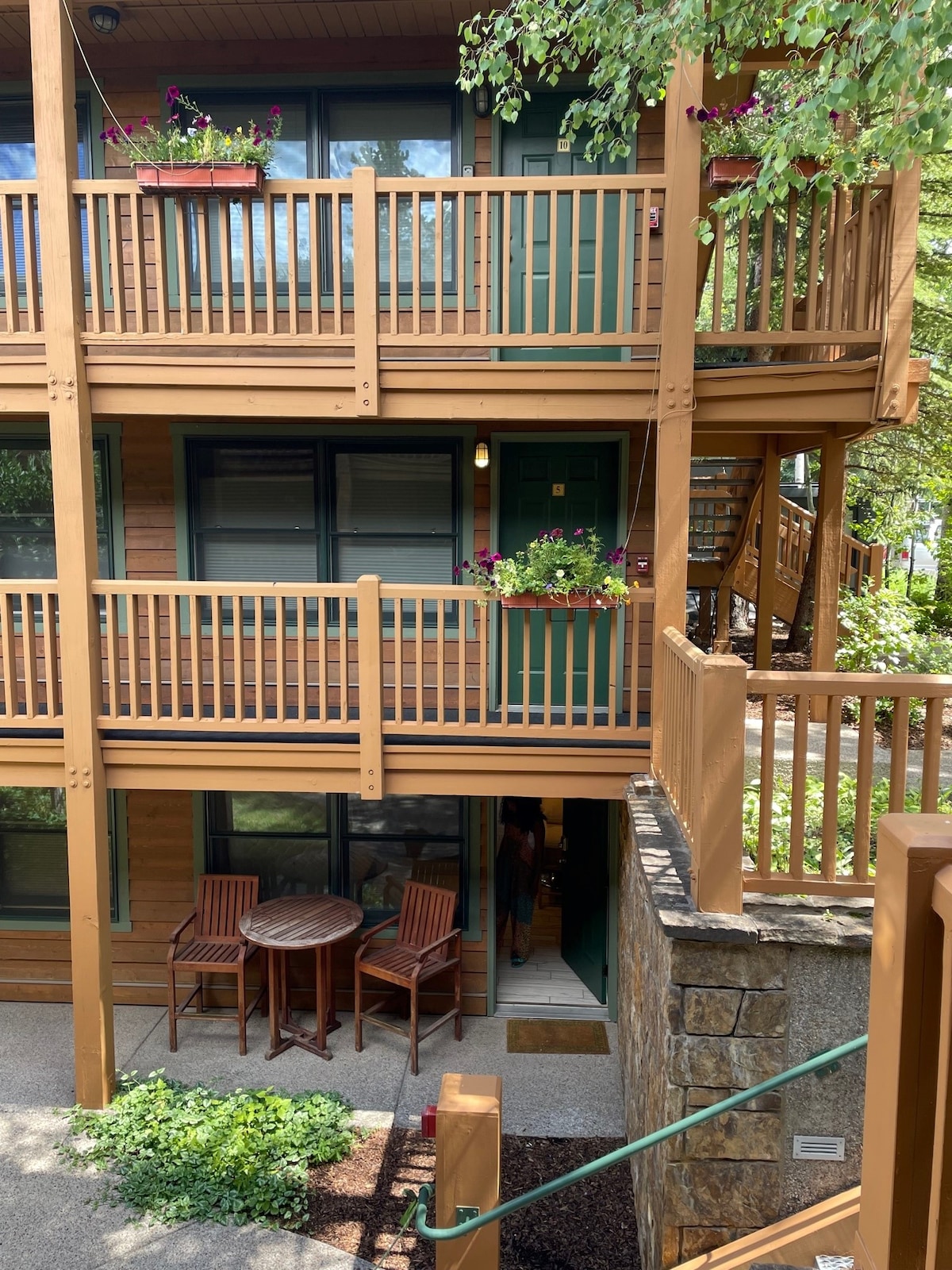 The exterior of the condo features a wooden balcony adorned with vibrant flower planters. A small round table with two chairs sits outside the entrance, surrounded by lush greenery. The inviting entrance is framed by large windows, highlighting the unit's connection to the outdoors.