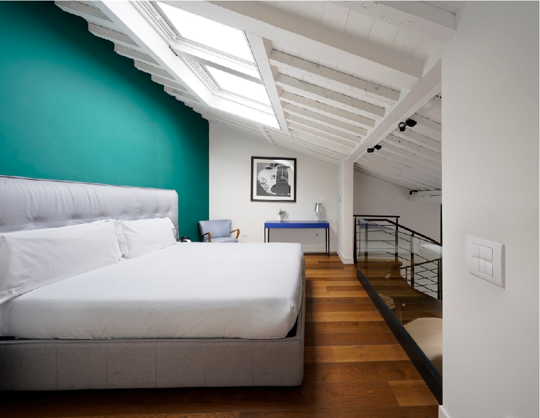 A spacious bedroom is presented with a large double bed against a dark green wall. Natural light enters through skylights in the sloped ceiling, illuminating the room's wood flooring. A modern chair and a side table with a lamp complete the inviting setup.