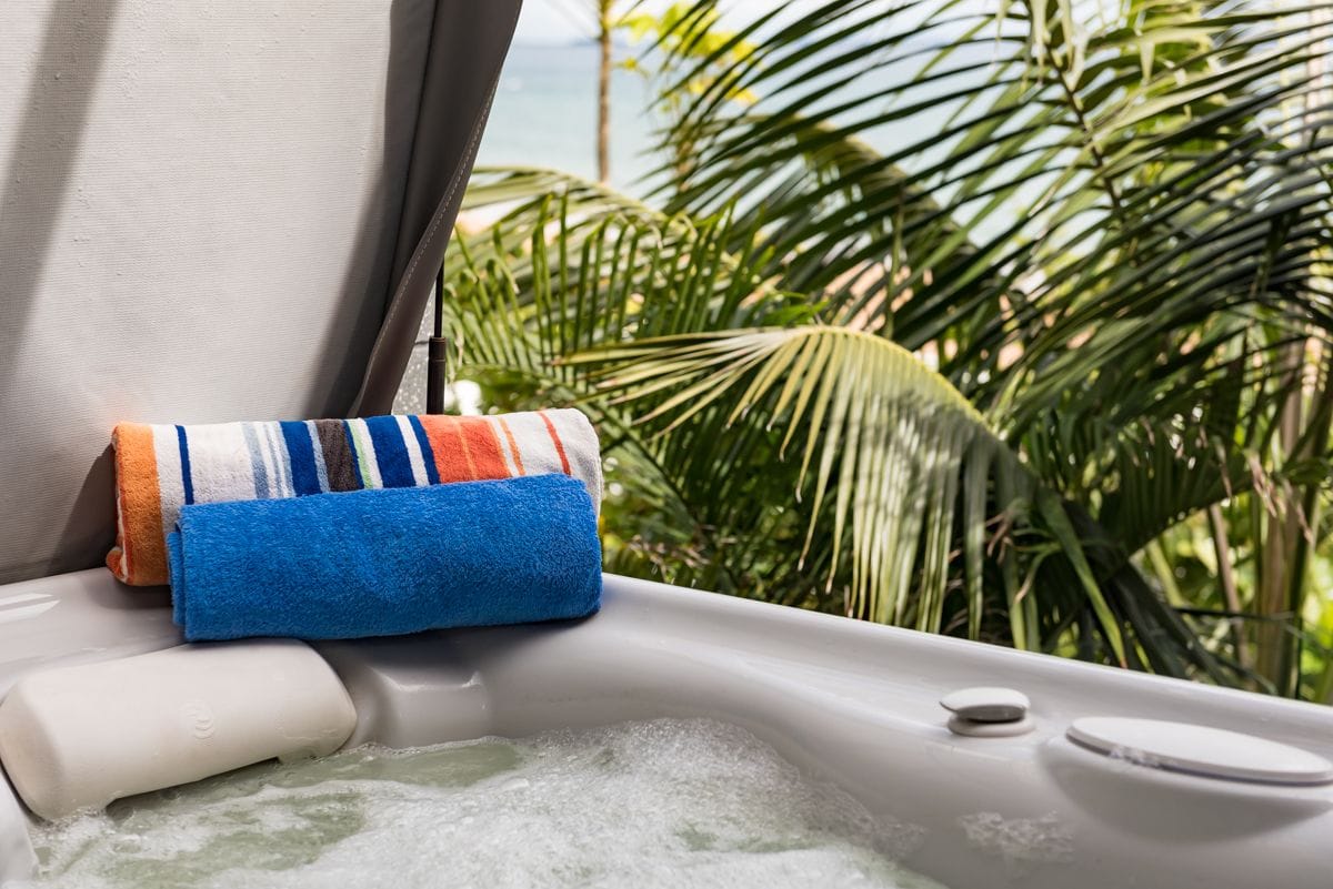 A spa bath is positioned on a shaded deck, with soft bubbling water visible. A neatly folded orange and blue striped towel rests beside the bath, surrounded by lush greenery. The calming view of the ocean can be seen in the background through the foliage.