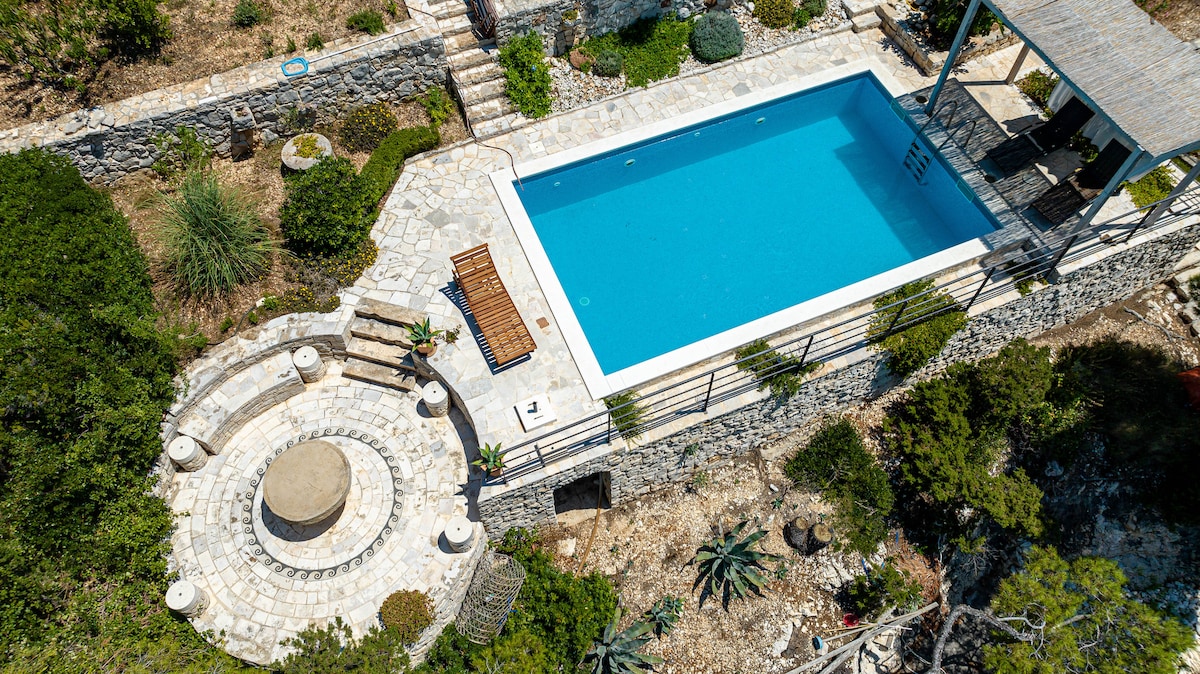 An aerial view showcases a serene pool surrounded by natural stone pathways and lush greenery. A spacious patio area features lounge chairs and a circular seating arrangement, creating a welcoming space for relaxation. The clear blue water of the pool invites tranquil moments under the sun.