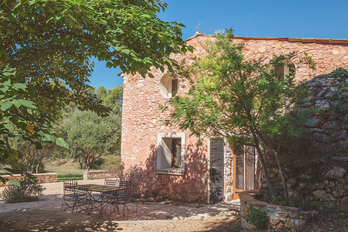 The rustic stone farmhouse features a blend of warm hues and natural textures. Sunlight filters through the surrounding trees, casting gentle shadows on the outdoor patio equipped with a metal dining set, perfectly situated to enjoy the serene landscape.