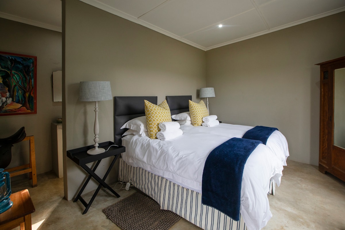 A spacious bedroom is furnished with two beds, each dressed in crisp white linens, complemented by yellow patterned pillows. Soft lighting is provided by lamps on either side, and a textured rug is placed beneath a small side table, enhancing the room's comfort.