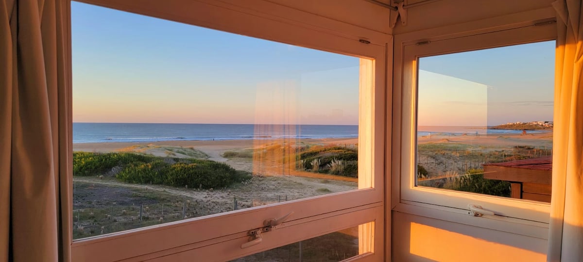 A view from the interior space captures the serene ocean landscape through large windows. The horizon showcases gentle waves and soft sandy dunes, illuminated by the warm colors of sunset. A calm atmosphere is reflected in this coastal scene.