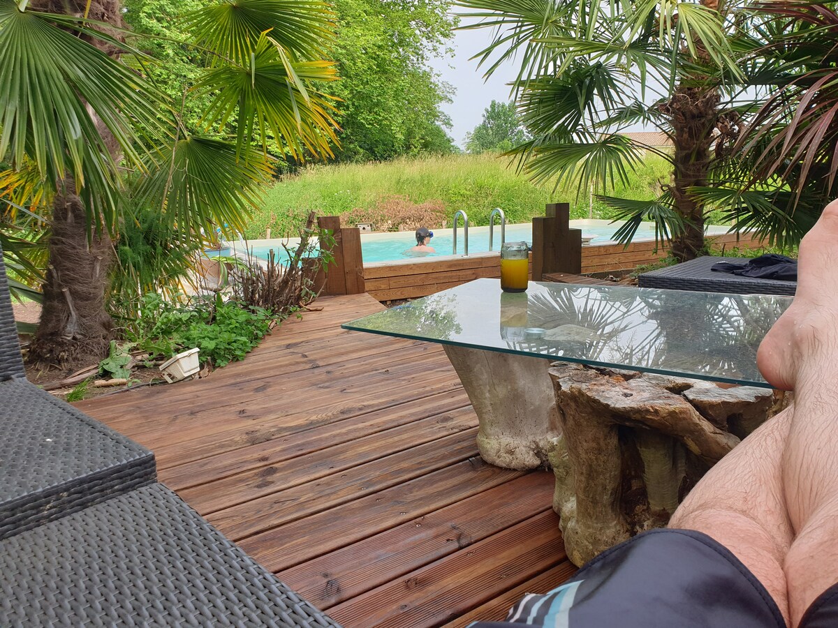 A peaceful outdoor space is showcased, featuring wooden decking with a glass table and stylish seating. Tropical plants surround the area, and a swimming pool is visible in the background. A figure can be seen enjoying the water, adding to the serene atmosphere.