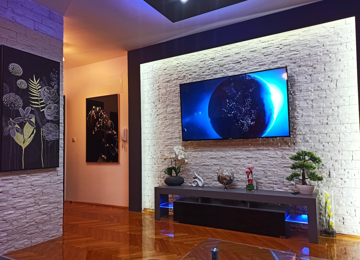 The living room features a modern design with a large flat-screen TV mounted on a textured wall. A low-profile console table holds decorative items and plants. Soft lighting enhances the room's ambiance, complemented by artwork displayed on adjacent walls.