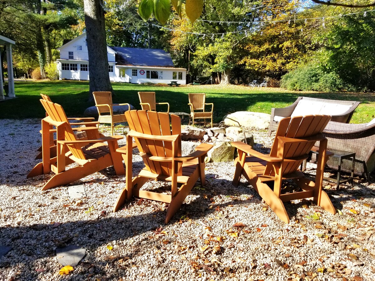 A tranquil outdoor seating area is highlighted by wooden adirondack chairs arranged around a stone fire pit. Lush greenery surrounds the space, complemented by a gravel surface. A backdrop of the country estate and a distant building enhances the inviting atmosphere.