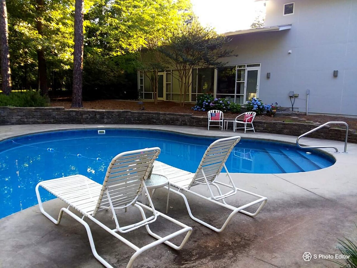 A refreshing salt-water pool is framed by a spacious deck, featuring two lounge chairs positioned for relaxation. The surrounding area is enhanced by greenery, with large windows visible on the nearby home, inviting natural light into the space.