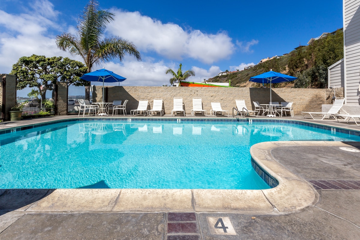A clear blue pool is surrounded by lounge chairs and umbrellas, creating a relaxing outdoor space. Lush palm trees can be seen in the background, while the calm sky adds to the serene ambiance.