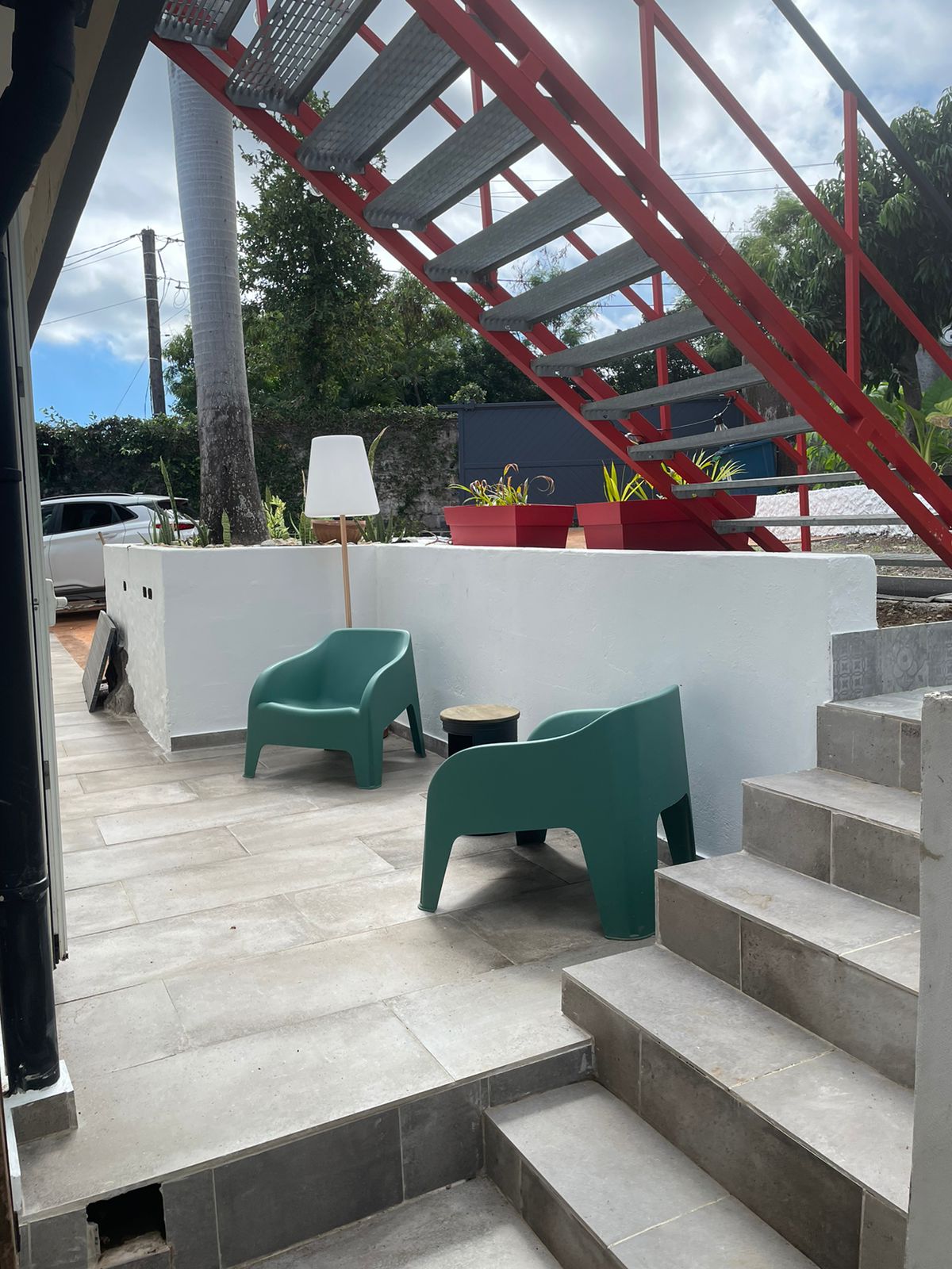 An outdoor area features two green chairs and a small table, surrounded by a simple wall and planters. A staircase with red railings leads to the upper level, while the flooring consists of light-colored tiles, complementing the natural setting.