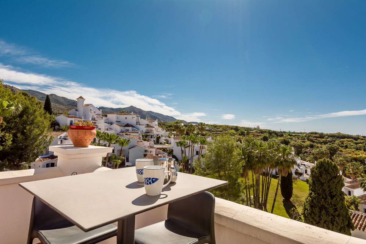 A spacious terrace is shown with a modern table and four chairs. Two cups and a small pot of flowers are placed on the table. The background reveals a scenic view of the surrounding tropical gardens and white buildings, framed by clear blue skies.