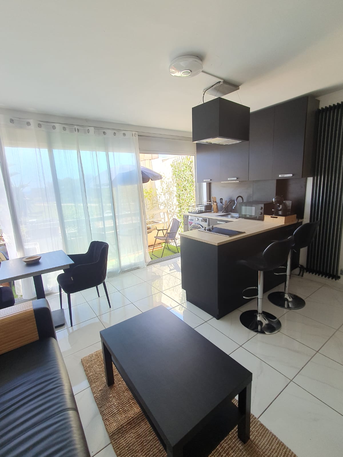 A modern kitchenette is equipped with a sleek island and bar stools, offering seating for two. Natural light filters through sheer curtains. A dining table for two is positioned nearby, complemented by a comfortable seating area featuring a dark couch and a decorative rug.
