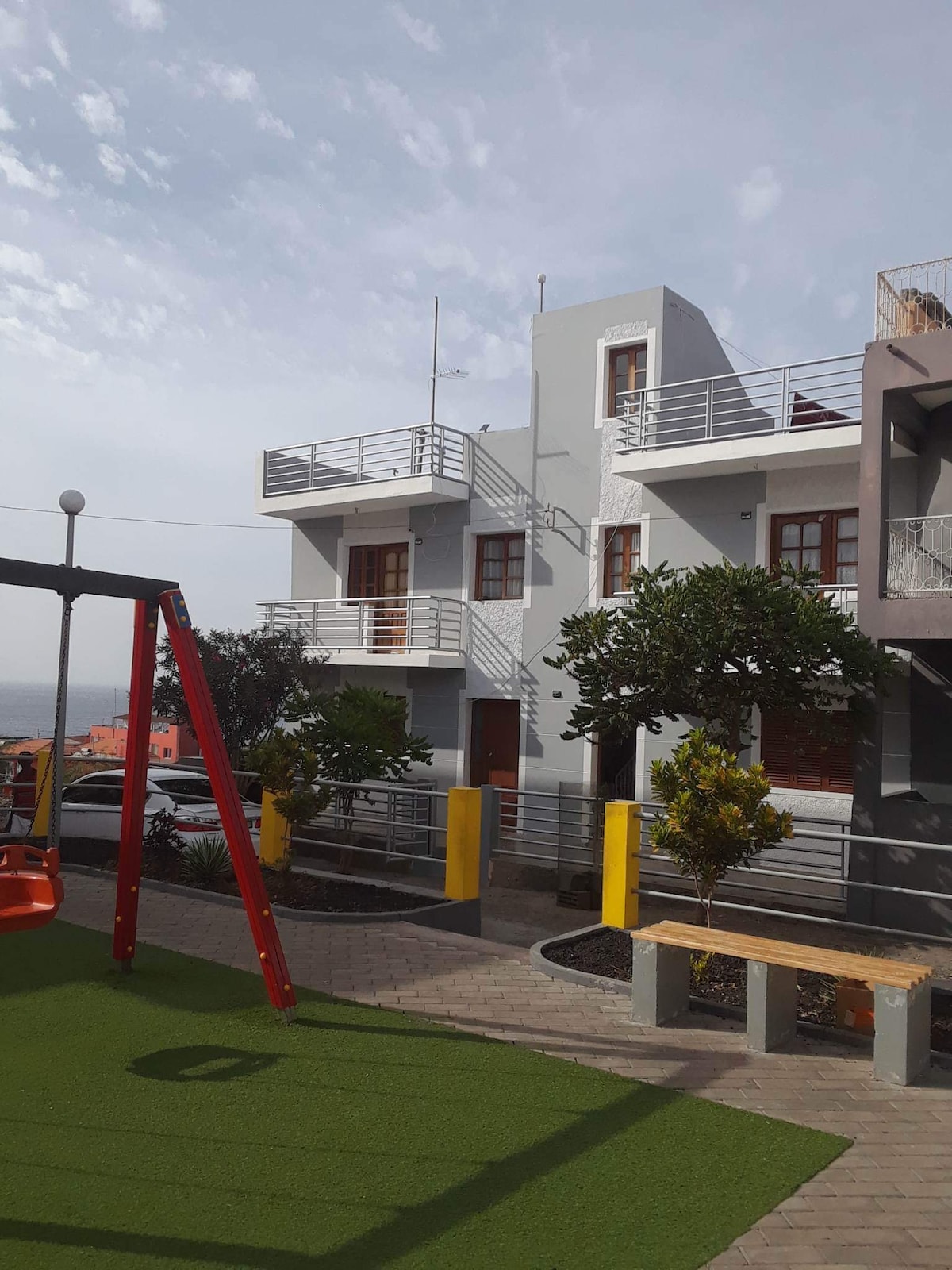 A multi-story building with large windows and balconies is shown. In the foreground, a playground area features a swing set against a backdrop of manicured greenery and landscaping. The scene is bright, revealing a peaceful environment with a glimpse of the surrounding area.