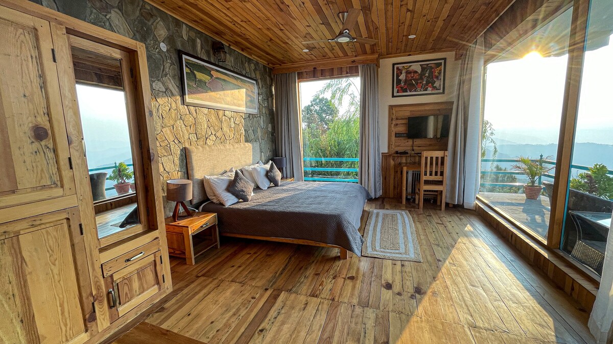 A spacious room features wooden flooring and walls adorned with natural stone. A large window offers breathtaking views of the surrounding mountains, while sunlight enhances the warm tones of the wooden furnishings. The room includes a cozy bed, a TV, and a small dining area.