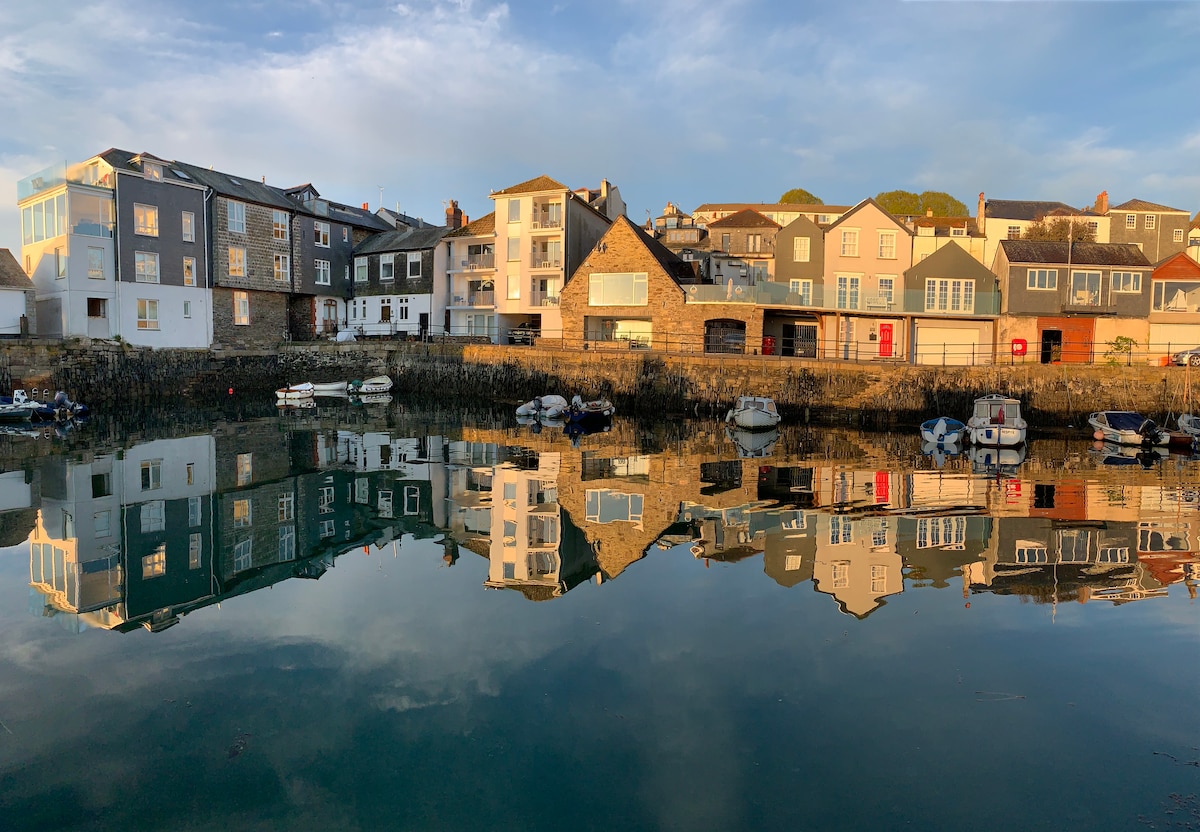 Waterfront buildings line the quayside, their reflections visible in the calm water. The architecture showcases a mix of styles, with various colors and window designs. Small boats are anchored along the shoreline, adding to the tranquility of the scene.
