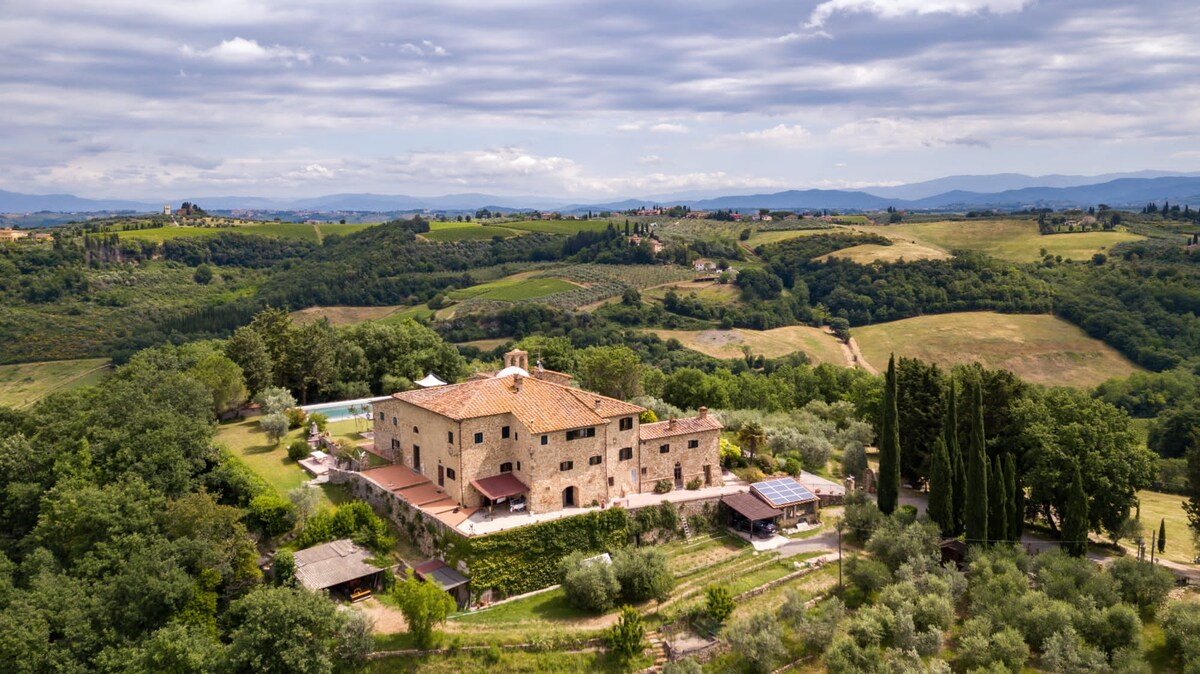 An aerial view reveals a charming stone villa surrounded by lush green hills and countryside. A sparkling pool glistens in the sunlight, while various trees and gardens enhance the serene landscape. The rolling hills stretch into the distance under a partly cloudy sky.